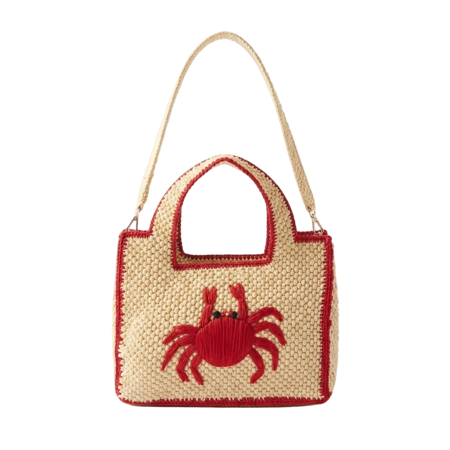 A woven straw handbag with a red border and a red embroidered crab on the front, featuring a light beige shoulder strap, set against a plain white background.