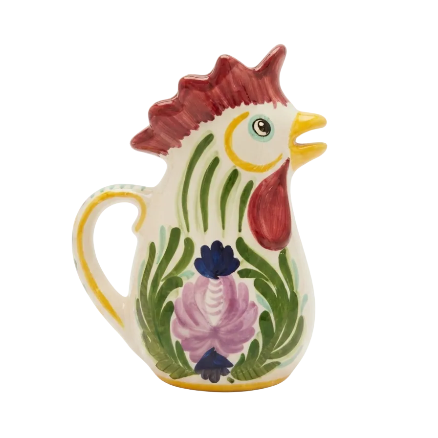 A ceramic pitcher shaped like a rooster, featuring a cream-colored body with hand-painted details of a red comb and wattle, green feathers, a pink flower, and a yellow beak and handle trim, set against a plain white background.