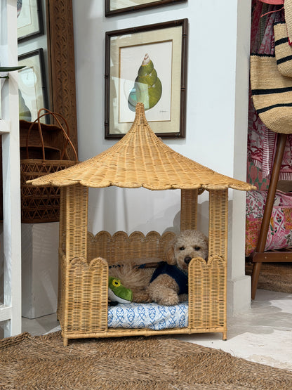 A small fluffy dog sits inside a woven pagoda shaped wicker pet bed with a thatched roof and blue patterned cushion.